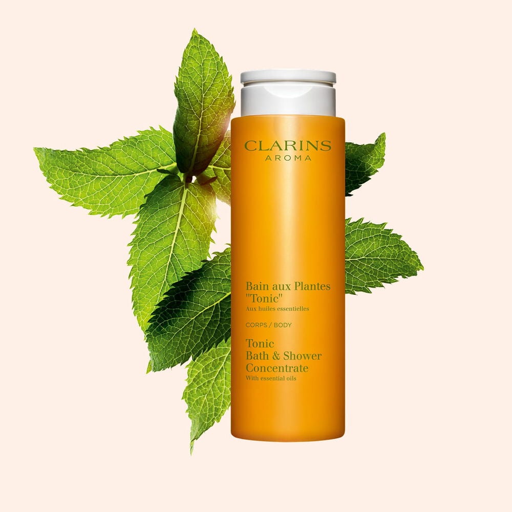 Packshot del Bagno Doccia Piante &ldquo;Tonicit&agrave;&rdquo; Clarins vicino a foglie di menta e rosmarino su uno sfondo arancione.