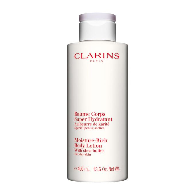 Packshot di un flacone di Crema Corpo Super Idratante Clarins vicino ad alcuni semi di karit&eacute; su uno sfondo bianco.