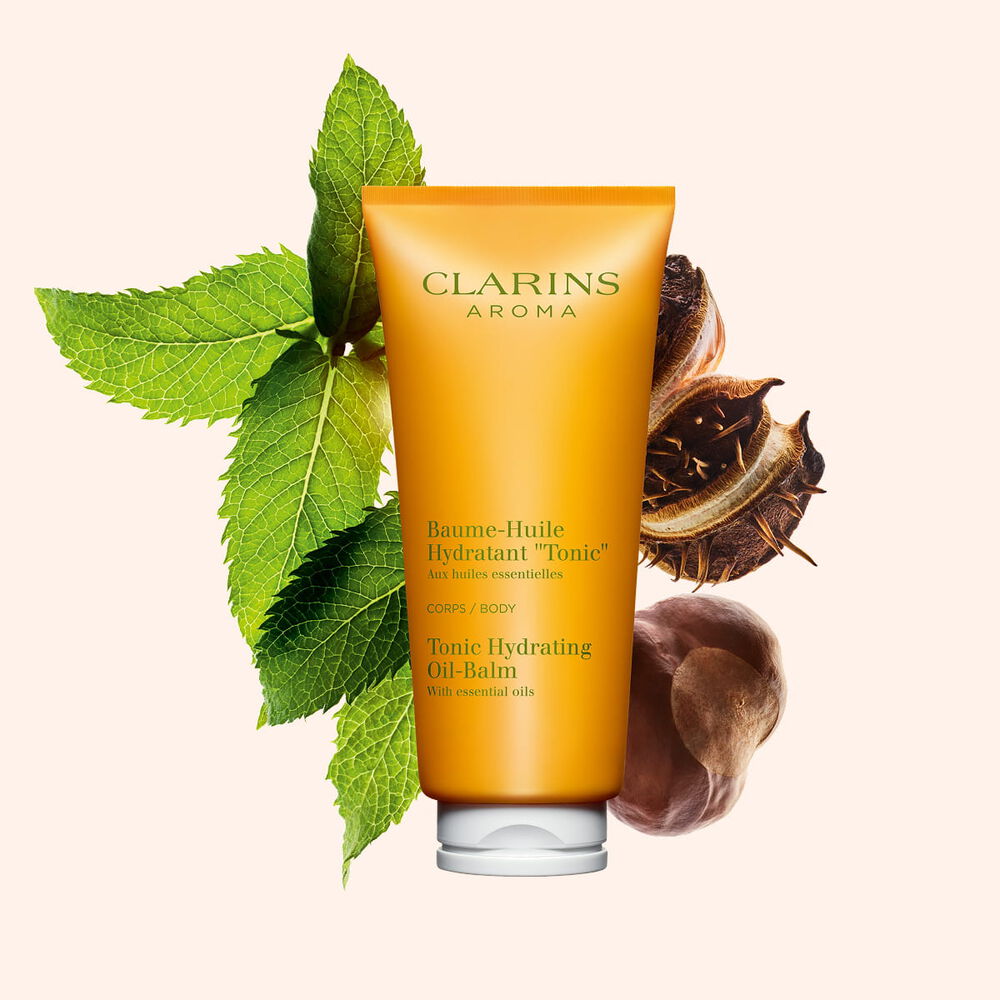 Packshot di un tubetto di Crema-in olio idratante corpo &ldquo;Tonicit&agrave;&rdquo; vicino a foglie di menta e semi di karit&eacute; su uno sfondo color pesca.