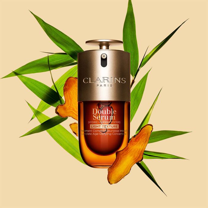 Un flacone da 30 ml di Double Serum Light Texture di Clarins tra curcuma e foglie di canna di Provenza su sfondo arancione.