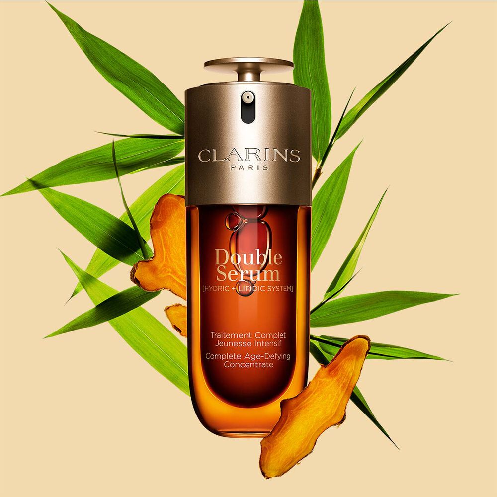 Packshot di Double Serum Clarins mostrato di fronte ai suoi ingredienti, tra cui la curcuma, su uno sfondo dorato
