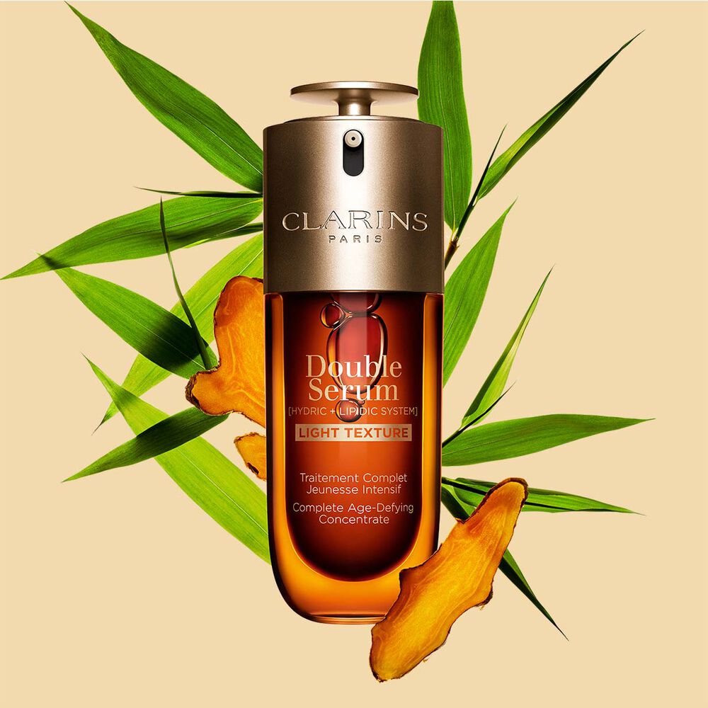 Un flacone da 70 ml di Double Serum Light Texture di Clarins tra curcuma e foglie di canna di Provenza su sfondo arancione.