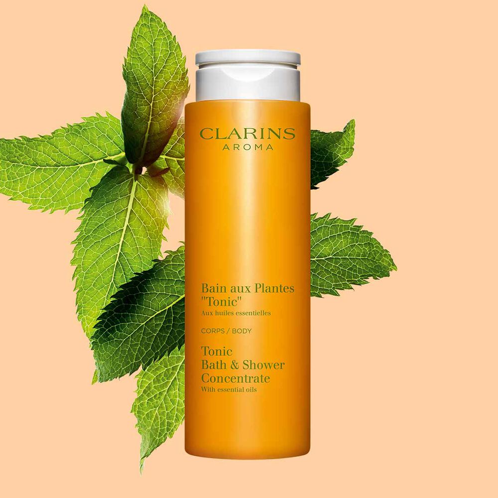 Packshot del Bagno Doccia Piante &ldquo;Tonicit&agrave;&rdquo; Clarins vicino a foglie di menta e rosmarino su uno sfondo arancione.