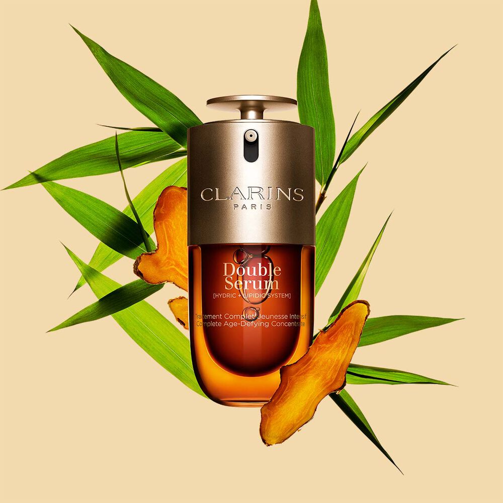 Packshot di Double Serum Clarins mostrato di fronte ai suoi ingredienti, tra cui la curcuma, su uno sfondo dorato