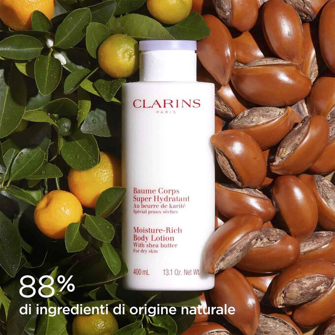 Flacone di Crema Corpo Super Idratante Clarins circondato da semi di karit&eacute; e arance per illustrare i suoi ingredienti naturali.